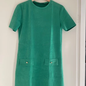 Grön klänning från Zara - Säljer en snygg grön klänning från Zara i storlek S. Den har korta ärmar och två dekorativa fickor framtill med guldknappar. Perfekt för vår och sommar! 💚
