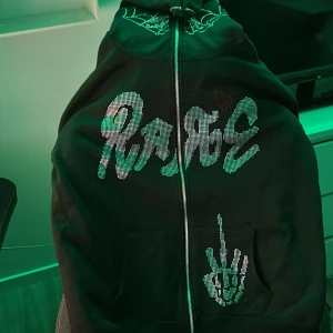Svart hoodie med tryck - Cool svart hoodie med dragkedja och tryck av texten 'RAGE' i grönt. Har en unik design med ett skelettmönster på framsidan. Perfekt för en avslappnad stil eller en kväll med vänner. Passar bra till jeans eller joggers.