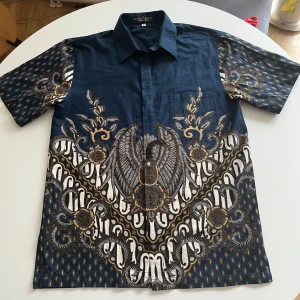Mönstrad batikskjorta från Bali - Säljer en unik batikskjorta i mörkblått med ett detaljerat mönster i vitt och guld. Skjortan har korta ärmar och en klassisk krage. Perfekt för sommaren. Materialet känns lätt och luftigt, vilket gör den perfekt för varma dagar. Inköpt på resa till Bali.