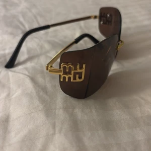 Bruna solglasögon med gulddetaljer - Snygga bruna solglasögon med guldfärgade detaljer och en unik design på sidorna. De har en modern look med stora glas och tunna skalmar. Perfekta för en solig dag och ger en lyxig känsla. Passar både till vardags och fest!