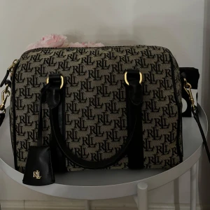 Mönstrad väska från Lauren Ralph Lauren - Snygg och stilren väska från Lauren Ralph Lauren med RLL-logga över hela ytan. Väskan är i ett mönstrat tyg med svarta detaljer och har en praktisk dragkedja. Perfekt för både vardag och fest!
