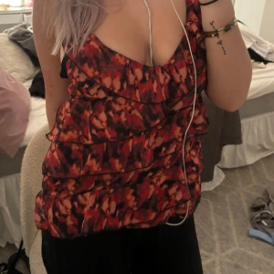 Mönstrad ärmlös topp - Säljer en snygg ärmlös topp med ett coolt mönster i rött, svart och orange. Toppen har volangdetaljer och en djup urringning, perfekt för festliga tillfällen. Den är i bra skick och passar perfekt till både jeans och kjol.