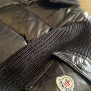Svart dunjacka från Moncler - Säljer en snygg svart dunjacka från Moncler. Jackan har en glansig finish med stickade detaljer och en dragkedja framtill. Perfekt för kyliga dagar när du vill hålla stilen. Moncler-loggan syns tydligt på ärmen. Passar både till vardags och lite finare tillfällen.