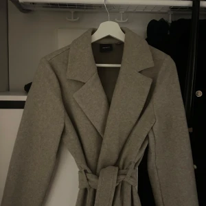 Beige kappa från Vero Moda - Säljer en snygg beige kappa från Vero Moda i storlek S. Den har en klassisk design med knytskärp i midjan och långa ärmar. Perfekt för höst och vår! 🧥
