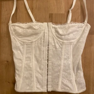 Vit spetskorsett med snörning - Säljer en vit korsett med vacker spetsdetalj och snörning i ryggen. Den har justerbara axelband, som passar perfekt för festliga tillfällen. Korsetten är i mycket bra skick och ger en fin passform.