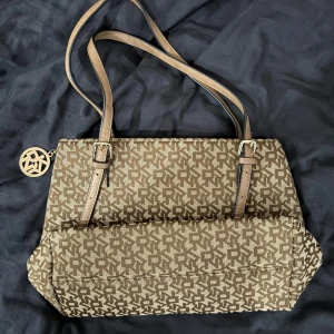 Beige och brun monogram axelväska  DKNY - Säljer en axelväska beige och brunt med ett coolt mönster. Insidan är rymlig med flera fack för bra organisering. Perfekt för både vardag och fest! Väskan är kanske använd en gång, har inga dpår av användning. Väskan är inte från varumärket DKNY