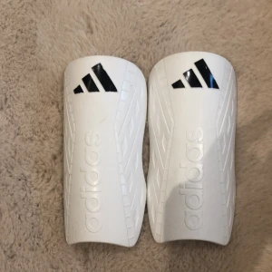 Vita benskydd från Adidas - Säljer ett par vita benskydd från Adidas i mycket bra skick. De har en stilren design med svarta detaljer och Adidas-loggan. Perfekta för fotbollsträningen eller matchen. Passar bra för både kvinnor och män. Storleken är för alla
