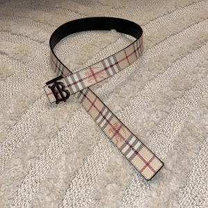 Burberry bälte - Säljer ett snyggt vändbart bälte burberry med ett klassiskt rutigt mönster i beige på ena sidan och stilrent svart på andra sidan. Bältet har ett coolt monogramspänne i silverfärg. Perfekt för både vardag och fest! 🖤(däremot lite skrapat på märket)