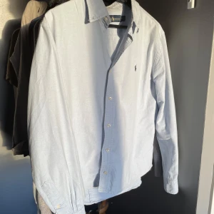 Ljusblå skjorta från Ralph Lauren - Säljer en stilren ljusblå skjorta från Ralph Lauren i storlek M. Skjortan har långa ärmar och klassiska knappar framtill. Den är i mycket bra skick och har det ikoniska broderade logotypen på bröstet. Perfekt för både vardag och fest! Det står att den är storlek M men misstänker att det är en dam model. Men satt riktigt bra på mig när jag var mellan 160/170 cm lång. Men säljs nu när den är för liten. Hör gärna av dig vid funderingar!  Mvh Rufus 