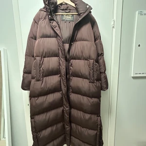 Brun lång dunjacka från Superdry - Säljer en brun lång dunjacka från Superdry i storlek EU 42. Jackan är perfekt för kalla vinterdagar med sin pufferstil och långa ärmar. Den har en praktisk huva och stängs med dragkedja. Superdry-loggan finns på insidan. Perfekt för att hålla dig varm och stilren i vinter, har själv haft den i minus 20 - 30 grader! 