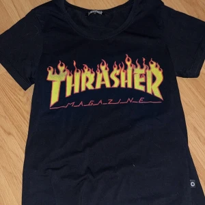 Svart Thrasher t-shirt - Säljer en svart Thrasher t-shirt i bomull med det ikoniska flammande logotyptrycket i gult och rött. Perfekt för en casual look eller skateparken. T-shirten är kortärmad och i mycket bra skick. Passar perfekt till jeans eller shorts!