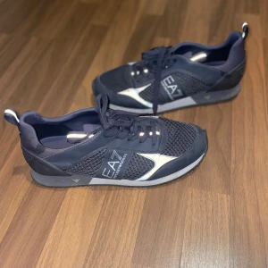 Svarta sneakers från EA7 - Säljer ett par snygga svarta sneakers från EA7 i mycket bra skick. De har en stilren design med vita detaljer och är perfekta för både vardag och träning. Skorna har snörning och en bekväm passform. Perfekta för dig som vill ha både stil och komfort! 🖤