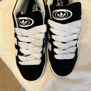 Svarta och vita sneakers från Adidas - Säljer ett par snygga Adidas Campus sneakers i svart och vitt. De har en klassisk design med vita ränder och en gummisula i brunt. Skorna är helt o använda och fräscha och är i storlek 44