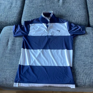 Randig pikétröja från Puma - Säljer en snygg randig pikétröja från Puma i blått och vitt. Tröjan har korta ärmar och en klassisk krage med knappar. Perfekt för både vardag och sportiga aktiviteter. Passar bra till jeans eller shorts för en avslappnad look. Pris går alltid att förhandlas!