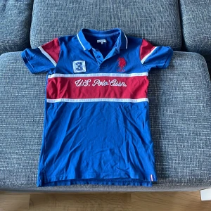 Blå och röd pikétröja från U.S. Polo Assn. - Säljer en snygg blå pikétröja från U.S. Polo Assn. med röda och vita detaljer. Tröjan har en klassisk krage och knappar framtill. Perfekt för en avslappnad men stilren look. Passar bra till både vardag och fest! Pris går alltid att förhandlas! Storlek 14/16 eller Xs 