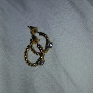 Guldörhängen med stenar - Snygga guldörhängen med en unik design. De har en cirkulär form med små kulor längs kanten och en glittrande sten i mitten. Perfekta för att ge lite extra bling till din outfit, oavsett om det är till vardags eller fest.