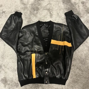 Svart skinnjacka från AMGE - Made in Spain - Säljer en snygg svart skinnjacka från AMGE, tillverkad i Spanien. Jackan har coola gula detaljer och knappar framtill. Perfekt för höst och vår med sin stilrena design och bekväma passform.