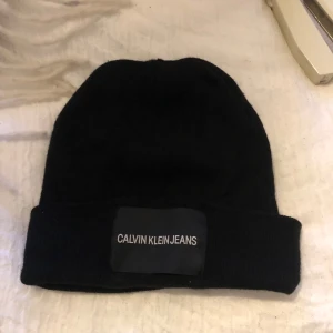Svart mössa från Calvin Klein - Snygg svart mössa från Calvin Klein Jeans i one size. Perfekt för att hålla sig varm under kyliga dagar. Den har en stilren design med en uppvikt kant och märkeslogga framtill. Passar både till vardags och mer avslappnade tillfällen.