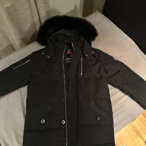 Svart parkajacka med pälsdetalj - Säljer en snygg svart parkajacka från Moose Knuckles. Jackan har en fluffig pälsdetalj på huvan och flera praktiska fickor med dragkedjor. Perfekt för kalla vinterdagar. Den är i mycket bra skick och redo för nya äventyr! 🖤