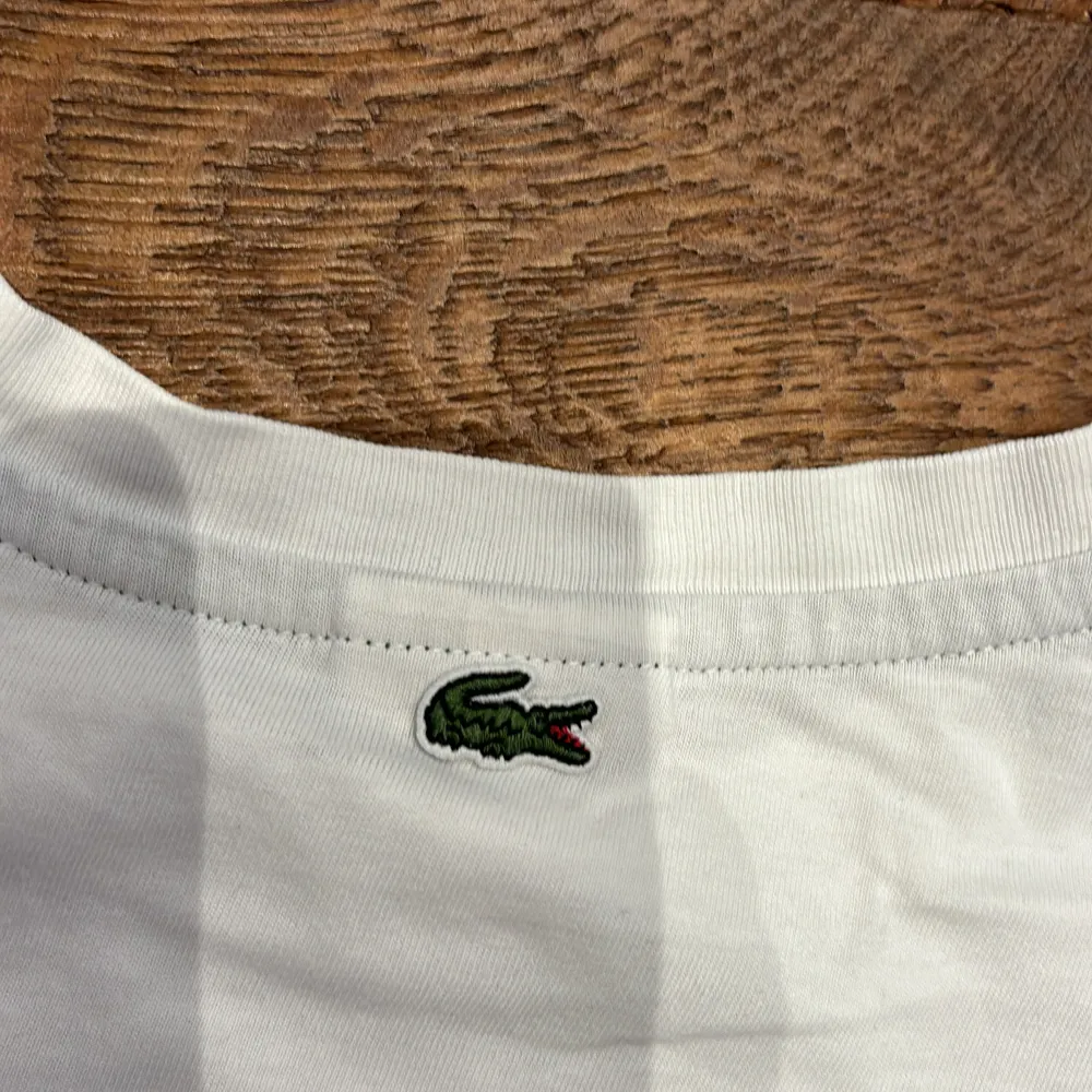 Säljer en klassisk vit t-shirt från Lacoste i storlek 14. Den har det ikoniska krokodilmärket broderat på framsidan och en grön rand vid kragen. Perfekt för en avslappnad stil eller sommarens äventyr. T-shirten är i bra skick och redo för en ny ägare!. T-shirts.