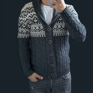 Mörkgrå stickad kofta med mönster - Säljer en snygg mörkgrå stickad kofta med ett stilrent vitt mönster upptill. Koftan har långa ärmar och knappar framtill, perfekt för kyliga dagar. Den är både varm och trendig, idealisk för höst och vinter. Passar bra till  jeans!