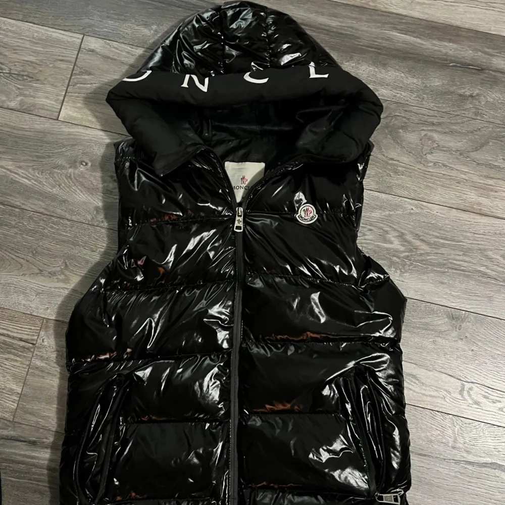 Säljer en snygg svart dunväst från Moncler i mycket bra skick. Västen har en glansig finish och en dragkedja framtill. Perfekt för höst och vinter, med en stor huva för extra värme. Moncler-loggan syns tydligt på bröstet och kragen.. Jackor.