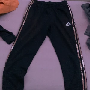 Svarta träningsbyxor från Adidas - Säljer ett par svarta träningsbyxor från Adidas med klassiska ränder längs sidorna. Byxorna har en elastisk midja och är perfekta för träning eller en avslappnad dag. De är i bra skick och redo för nya äventyr!