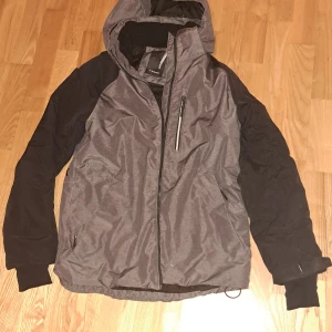 Grå och svart jacka - Säljer en grå och svart jacka med dragkedja. Den har långa ärmar och en praktisk ficka på bröstet. Perfekt för vinterns skidåkning. Lite slitage på ena ärmen, men annars i bra skick.