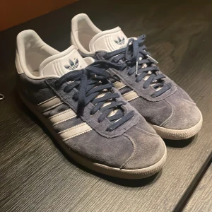 Adidas Gazelle i blå mocka - Säljer ett par klassiska Adidas Gazelle i blå mocka. De är i bra skick med lite slitage på sulorna och tappat lite färg på mockan. Har smuts aom man kan tvätta bort men fortfarande superbekväma och riktigt snygga. Inköpta för 1200kr. Pris går att diskutera vid snabb affär, Mvh Axel.