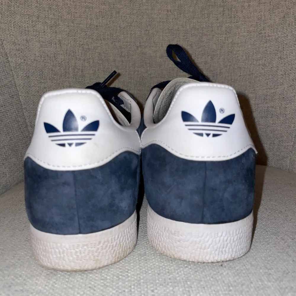 Säljer ett par snygga Adidas Gazelle i marinblå med vita detaljer. Skorna har den klassiska tre-randiga designen och är i mycket bra skick. Perfekta för både vardag och fest! Skorna har en vit sula och snörning i matchande blå färg. Priset går att sänkas!!. Skor.