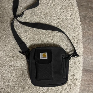 Svart Carhartt essentials bag - Stilren och praktisk svart axelväska från Carhartt. Väskan har en justerbar axelrem och flera fack med dragkedja. Säljer då jag aldrig använder den mer 💕