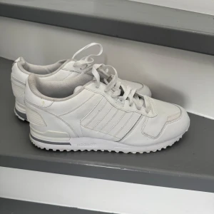 Vita sneakers från Adidas - Säljer ett par vita Adidas sneakers i bra skick. De har klassisk design med snörning och den ikoniska trefoil-loggan på hälen. Perfekta för vardagsbruk och passar till det mesta. Sulan är i bra skick och de är bekväma att gå i. Perfekta för både vår och sommar!
