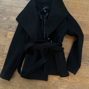 Svart kappa från vero Moda  - Snygg svart kappa med stort krage och bälte i midjan. Perfekt för höst och vår. Den har en elegant och tidlös design som passar både till vardags och fest. Kappan är i bra skick och har långa ärmar.