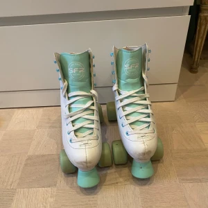 Vita rullskridskor från SFR - Säljer ett par vita rullskridskor från SFR i storlek EU 38. De har ljusblå detaljer och snörning. Perfekta för fritidsåkning! Skorna är i bra skick med några mindre märken på tån. Hjul och broms är i ljusblått och ger en retro känsla. Passar perfekt för en rolig dag i parken eller på rinken!