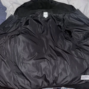 Svart pufferjacka från Hugo Boss - Säljer en snygg svart pufferjacka från Hugo Boss i storlek M. Jackan är perfekt för kalla dagar med sin vadderade design och huva. Den har en stilren look med diskret logga på insidan. Passar både till vardags och lite finare tillfällen. Håll dig varm och trendig i vinter! 