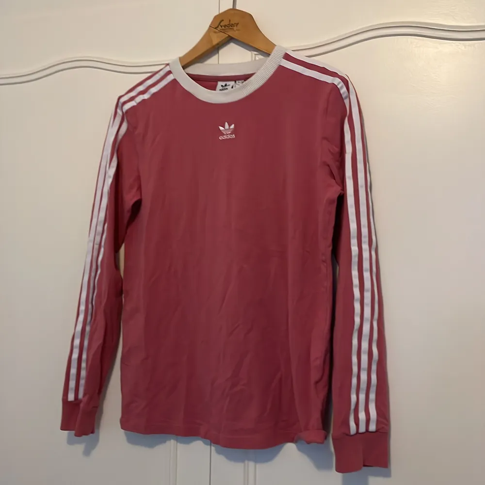 Säljer en snygg rosa långärmad tröja från Adidas med de klassiska vita ränderna längs ärmarna. Tröjan har en vit ribbad krage och är perfekt för en sportig look. Den är i bra skick och passar perfekt till både vardag och träning. 🌸. T-shirts.