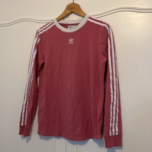 Rosa långärmad tröja från Adidas - Säljer en snygg rosa långärmad tröja från Adidas med de klassiska vita ränderna längs ärmarna. Tröjan har en vit ribbad krage och är perfekt för en sportig look. Den är i bra skick och passar perfekt till både vardag och träning. 🌸