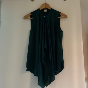 Mörkgrön ärmlös blus - Snygg mörkgrön blus i ett mjukt material. Den är ärmlös och har en elegant drapering framtill som ger en stilren look. Perfekt för både vardag och festliga tillfällen. Passar bra till både jeans och kjol!