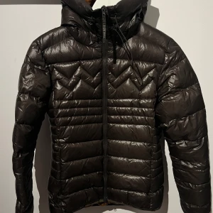 Brun dunjacka med huva - Säljer en snygg brun dunjacka från märket mountain works i gott skick. Jackan har en dragkedja framtill och en praktisk huva. Perfekt för kyliga höst- och vinterdagar. Den är långärmad och har en quiltad design som ger extra värme och stil. Passar både till vardags och lite mer uppklädda tillfällen. (Bak på armen finns ett litet hål, men inget som syns. Vi snackar inte ens 1 cm) 