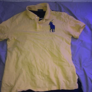 Gul och marinblå pikétröja från Polo Ralph Lauren - Säljer en snygg gul pikétröja från Polo Ralph Lauren med en stor marinblå logga på bröstet och siffran 3 på ryggen. Perfekt för en avslappnad stil eller en dag på stan. Tröjan är i bra skick och har korta ärmar. Passar perfekt till både jeans och shorts!