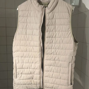Beige quiltad väst - Säljer en beige quiltad väst i bra skick. Den har en dragkedja framtill och är perfekt för höst och vår. Västen är stilren och passar till många olika outfits. Det finns en liten fläck på framsidan, men den är knappt märkbar. 