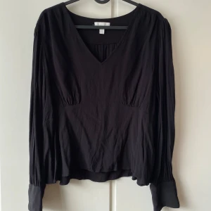 Svart blus med v-ringning - Snygg svart blus med v-ringning och långa ärmar. Perfekt för både vardag och fest. Den har en lös passform och är tillverkad i ett mjukt material som faller fint. Ärmsluten har knappar för en elegant touch.