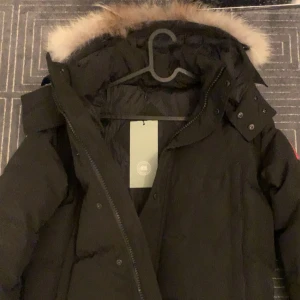 Svart dunjacka från Canada Goose - Säljer en svart dunjacka från Canada Goose i mycket bra skick. Jackan har en avtagbar huva med pälsdetalj och stängs med dragkedja och knappar. Perfekt för kalla vinterdagar! 🖤