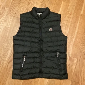 Svart dunväst från Moncler - Säljer en snygg svart dunväst från Moncler i mycket bra skick. Västen har en quiltad design med dragkedja framtill och två praktiska fickor med dragkedjor. Perfekt för höst och vår när du vill hålla dig varm och stilren. Passar ungdomar i storlek 152-158. Men har dok 2 hål men synat knappt!!!