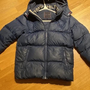Blå pufferjacka från Tommy Hilfiger - Säljer en blå pufferjacka från Tommy Hilfiger i andvänt skick. Jackan är i storlek 164 cm/ XS. Den har några små märken, och dragkedjan har blivit sliten. Därför säljer jag den för otroligt bra pris. Ny pris ca: 1500. Mitt pris:200