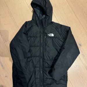 Svart dunjacka från The North Face - Säljer en svart dunjacka från The North Face. Den är perfekt för kalla dagar med sin quiltade design och långa ärmar. Jackan har en praktisk dragkedja och huva. Det finns en liten reva vid ärmen, men annars i bra skick. Perfekt för vintern! Jackan är vändbar så du kan välja mellan grå och svart. När du väljer den gråa färgen syns inte revan, inte heller den  lilla början till hål som uppträtt vid ärmslut på den svarta sidan. Vilket enkelt kan sys ihop. Jackan är för 13-14 år. 