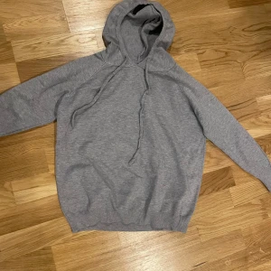 Grå hoodie - Säljer en grå hoodie i oanvänt skick. Passformen är normal och den har ribbade muddar vid ärmslut och nederkant.