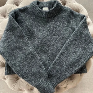 Grå stickad tröja - Mysig grå stickad tröja perfekt för kyliga dagar. Den har en klassisk design med ribbade muddar och en hög krage. Tröjan är i bra skick men har en liten reva som syns på bilden. Perfekt för höst och vinter!