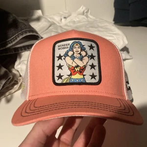 Rosa Wonder Woman keps - Snygg rosa keps med Wonder Woman-motiv framtill. Kepsen har ett broderat märke med stjärnor och Wonder Woman i mitten. Baksidan är i vitt meshmaterial med justerbart spänne för perfekt passform. Perfekt för sommarens äventyr eller som en cool accessoar till vardagsoutfiten. 10/10 i skick