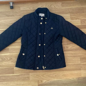 Mörkblå quiltad jacka från Hampton Republic - Säljer en snygg mörkblå quiltad jacka från Hampton Republic. Den har guldiga knappar och en broderad detalj med siffran 27 och en fågel. Perfekt för höst och vår, med en stilren look och normal passform. Jackan är i mycket bra skick och redo för nya äventyr! Vid frågor eller funderingar är du välkommen att meddela mig. Pris går diskuteras!
